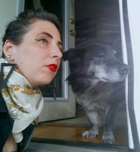 NB femme with bright red lipstick wearing triangle earings and a scarf and her black wolf-dog with very short legs looking out into the distance in a 3/4 view